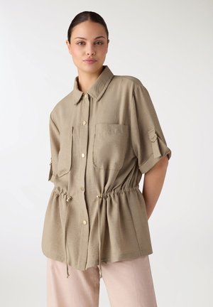 Giovane donna che indossa una camicia beige a maniche corte con bottoni, tasche sul petto e coulisse regolabili in vita, abbinata a pantaloni rosa chiaro.