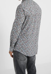 Camicia floreale a maniche lunghe con sfondo bianco, caratterizzata da piccoli fiori blu, rossi e verdi; polsini con bottoni e orlo curvo.