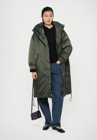 Manteau matelassé long de couleur vert olive avec un col haut, une fermeture éclair et un bouton, associé à un jean sombre et des ballerines noires. Petit sac noir.