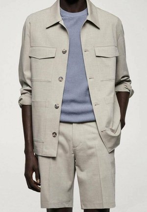 Mann trägt beige Jacke mit Knöpfen und Klappentaschen, passende Shorts und darunter einen blau strukturierten Pullover. Eine Hand in der Tasche, schlichter Hintergrund.