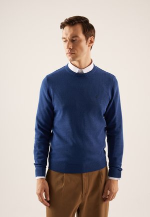 Blauwe gebreide trui met ronde halslijn en geribde manchetten, gecombineerd met een witte overhemd met kraag, gedragen over beige broeken.
