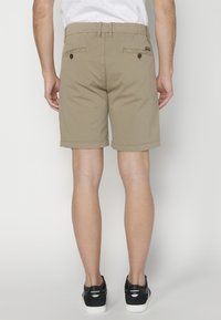 Vīrietis, tērpies beigtās šortos un melnos sporta apavos, stāv ar vēderu pie kameras pret vienkāršu fona.