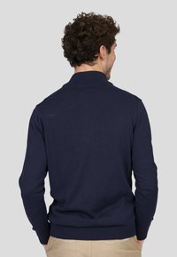 Pull en maille bleu marine avec un col montant, des bords-côtes aux poignets et à la taille. Le dos présente une texture lisse et une coupe ajustée.