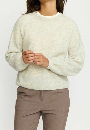 Femme portant un pull duveteux beige clair et un pantalon marron, debout les mains doucement jointes devant elle.