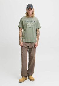 Grøn t-shirt med "ORIGINALS" tryk, brune løse bukser og gule støvler. Inkluderer en mørk kasket og synlige tatoveringer på armene.