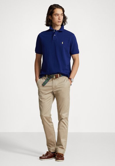 Moška modra polo majica s ovratnikom, v kombinaciji s bež chinos hlačami in rjavimi čolnarskimi čeveljci. Outfit vsebuje večbarvni zeleni pas.