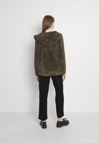 Oliven grøn, fuzzy jakke med en stor hætte, der har en tekstur, der ligner blødt fleece. Båret med sorte kordfløjspants og sko.