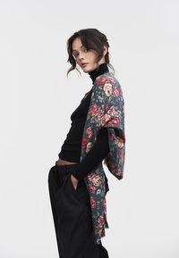 Mujer con cabello oscuro, vestida con un cuello alto negro y pantalones, posa con las manos en los bolsillos, llevando un chal azul con estampado floral sobre los hombros.