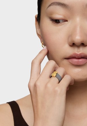 Dos anillos: un pendiente de aro de oro y un anillo de malla plateada con un acento de flor amarilla. La mano de la persona toca su cara. Fondo neutro.