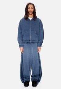 Άτομο με μακριά dreadlocks που φοράει μια ξεθωριασμένη μπλε ζακέτα με φερμουάρ και αντίστοιχο φαρδύ παντελόνι με ρίγες στα πλάγια, στέκεται μπροστά σε λευκό φόντο.