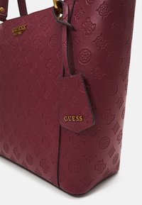 Borsa in pelle bordeaux con loghi impressi e dettagli in tonalità oro del marchio Guess sul davanti e un'etichetta attaccata.