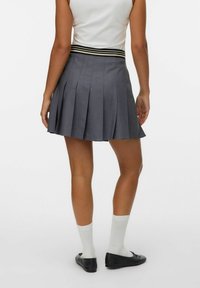 Femme portant un haut blanc et une jupe plissée grise avec une ceinture rayée, des chaussettes blanches et des ballerines noires, se tenant de dos sur un fond blanc.