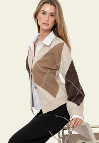 Jeune femme aux longs cheveux blonds portant un pull à motifs losanges marron clair et foncé sur une chemise blanche, pantalon noir, tenant des lunettes à monture écaille, assise sur une chaise.