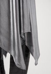 Un tissu métallique argenté drape avec une texture lisse et brillante ; il présente un ourlet asymétrique et tombe sur un matériau texturé noir en dessous.