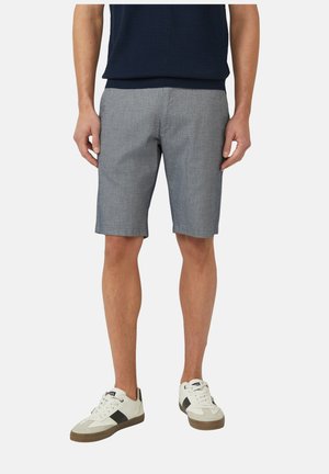 Mężczyzna w szarych szortach do kolan, granatowej koszuli i białych trampkach z czarnymi i brązowymi akcentami, stojący z luźno opuszczonymi ramionami.