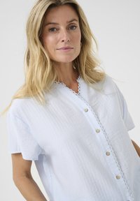 Camicia azzurro chiaro a maniche corte con bottoni, realizzata in tessuto testurizzato. Presenta un colletto con pizzo e bottoni in legno lungo la parte anteriore.