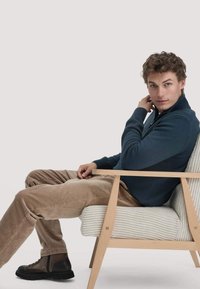 Donkerblauwe ritsvest, beige corduroy broek, bruine laarzen, zittend in een lichtgekleurde gestreepte fauteuil met houten frame.