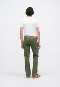 Mann som står med ryggen til, iført hvit t-skjorte, olivengrønne jeans med baklommer, og brune joggesko med beige detaljer.