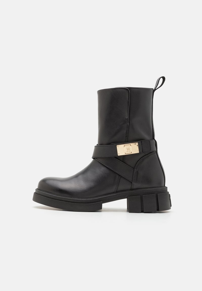 Botte en cuir noir montant jusqu'au milieu du mollet, avec une semelle épaisse, une languette à l'arrière et une sangle décorative avec boucle en métal doré sur le côté.