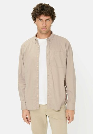 Manden med krøllet hår bærer en beige skjorte med knapper over en hvid t-shirt og beige bukser, stående mod en ensfarvet lys baggrund.