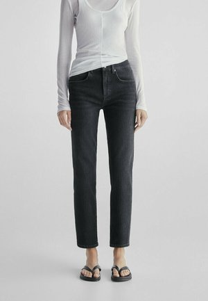 Kobieta ubrana w dopasowane czarne dżinsy, przezroczysty biały top z długim rękawem i czarne sandały z odkrytymi palcami, stoi na jednolitym jasnym tle.