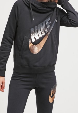 Osoba ubrana w czarną bluzę z kapturem Nike oraz dopasowane czarne spodnie z dużymi metalicznymi miedzianymi i srebrnymi logo Nike na obu elementach.