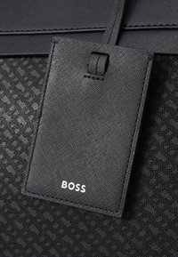 Juoda tekstūruota rankinė, pasižyminti lygiu audiniu ir raštuota šonine dalimi, kartu su nuimamu bagažo žymekliu, išrašytu „BOSS“. 