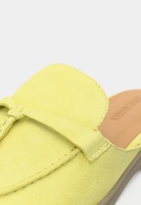 Mocasín sin cordones de ante amarillo con detalles cosidos y una puntera cuadrada redondeada, sobre una suela marrón claro.