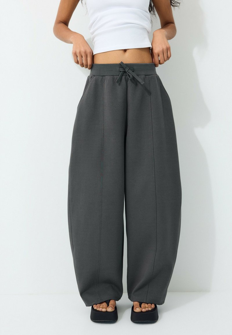 Person iført mørkegrå vidde bukser med snor i taljen, hvid crop top og sorte platformsandaler, stående mod en hvid baggrund.