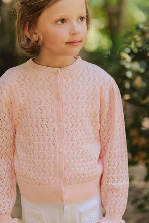 Lichtroze gebreide cardigan met een textuurpatroon, geribbelde manchetten en vijf witte knopen aan de voorkant. Draagt over witte broeken.