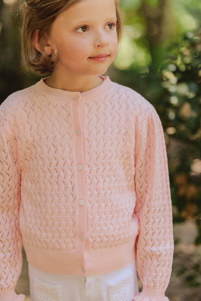 Helles rosa, gestricktes Cardigan mit einem strukturierten Muster, gerippten Manschetten und fünf weißen Knöpfen auf der Vorderseite. Über weißen Hosen getragen.