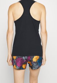 Nainen, jolla on päällä musta urheilullinen toppi ja kukkashortsit, joissa on suuria purppuran, keltaisen ja sinisen värisiä kukkia, nähtynä selkäpuolelta.