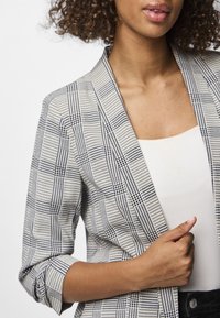 Blazer aos quadrados cinza e branco com colarinho entalhado, mangas dobradas e textura ligeira; usado por cima de um top claro, com decote natural.