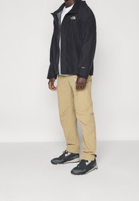 Černá voděodolná bunda, béžové kalhoty, černé tenisky. Bunda má zip vpředu, síťovou podšívku a logo značky na hrudi; kalhoty mají boční kapsy.