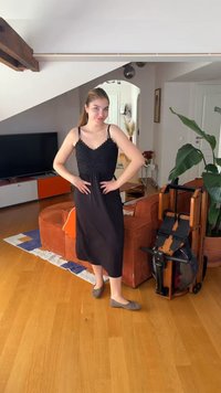 Jeune femme en robe noire posant avec les mains sur les hanches dans un salon avec un sol en bois, un canapé orange, une télévision et du matériel d'exercice.