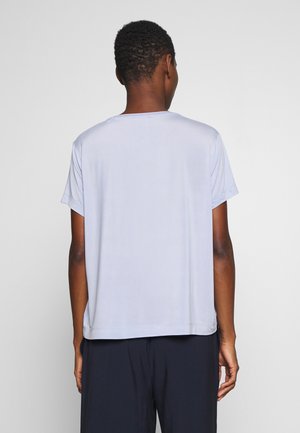 Osoba widziana od tyłu, ubrana w prostą, jasnoszarą koszulkę z krótkim rękawem oraz ciemne spodnie na jednolitym jasnym tle.