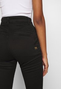 Pantalones negros de alta cintura hechos de tela duradera, con bolsillos traseros y una pequeña etiqueta de marca amarilla en el lado derecho.