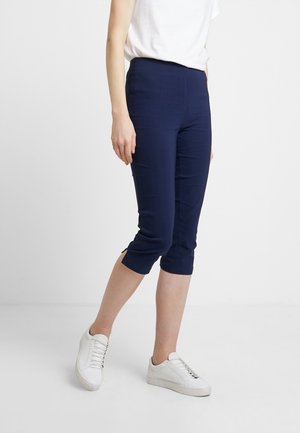 Persoon die marineblauwe cropped broek en witte sneakers draagt, staand met één been iets naar voren op een effen witte achtergrond.