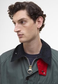 Πράσινο μπουφάν με κολάρο από κοτλέ σε μαύρο χρώμα και κόκκινη λεπτομέρεια στα άκρα, διαθέτει χρυσό φερμουάρ και κουμπί κλεισίματος. Το ύφασμα είναι λείο και υφή.