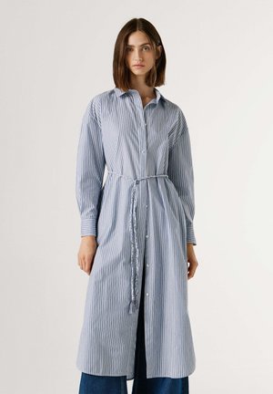 Donna che indossa un abito camicia lungo a righe verticali blu e bianche con bottoni e cintura intrecciata, in piedi davanti a uno sfondo chiaro e semplice.