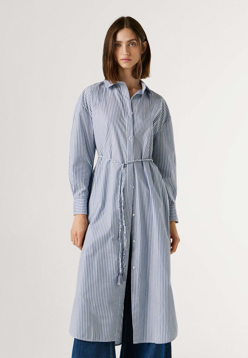 Γυναίκα που φοράει ένα μακρύ φόρεμα πουκάμισο με κάθετες μπλε και λευκές ρίγες, κουμπιά και πλεκτή ζώνη, στέκεται μπροστά σε ένα απλό ανοιχτόχρωμο φόντο.