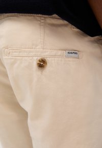 Chino bej cu o textură netedă, dotat cu un singur nasture, buzunar pe spate și o etichetă albă inscripționată "SCALPERS."