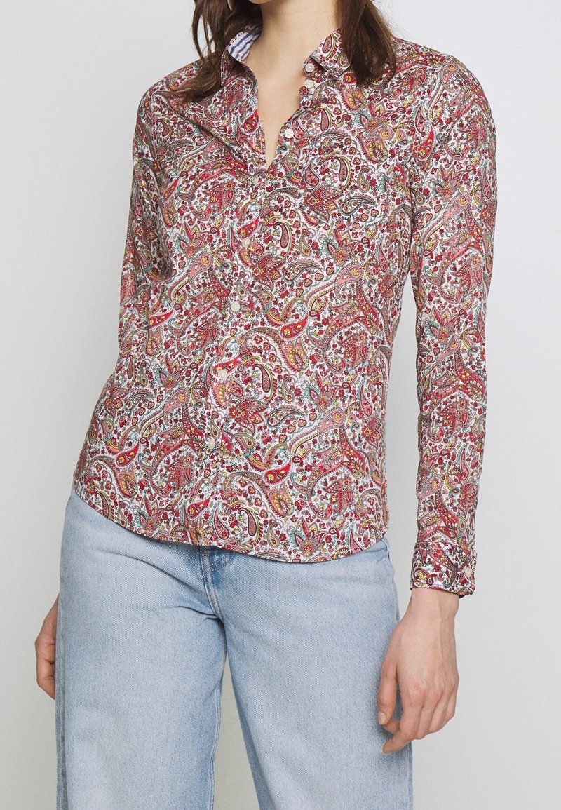 Femme portant une chemise boutonnée à manches longues avec un motif cachemire rouge et un jean bleu clair, debout devant un fond clair uni.