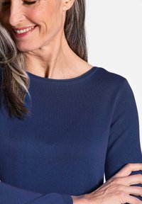 Marineblaues, geripptes Langarmoberteil mit Rundhalsausschnitt. Weiche Textur, taillierte Passform, die das Schlüsselbein und den Arm betont, ohne zusätzliche Details.