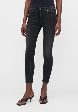 Kvinde iført sorte skinny jeans og sorte sandaler med hæl, stående foran en ensfarvet lys baggrund.