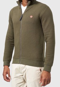 Veste zippée en tissu vert olive avec un motif texturé et matelassé. Comprend des poignets et un ourlet côtelés, ainsi qu'un petit patch logo sur la poitrine.