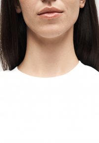 Κάτω μέρος προσώπου και λαιμός ενός ατόμου με μακριά σκούρα μαλλιά που φοράει λευκή μπλούζα, με τα χείλη κλειστά και νευρική έκφραση, σε λευκό φόντο.