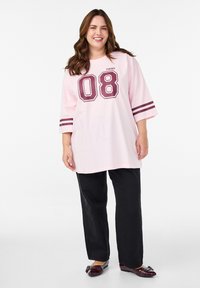 Ružové nadrozmerné tričko s vínovým nápisom "08" a "PARIS", s krátkymi rukávmi ozdobenými dvomi bielymi pruhmi. Dopĺňa ho čierne široké nohavice.