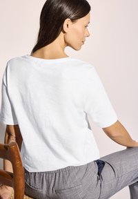 Witte t-shirt met korte mouwen en een relaxte pasvorm, met een ronde halslijn. Draag het met grijze gebreide broek, zittend tegen een lichte achtergrond.
