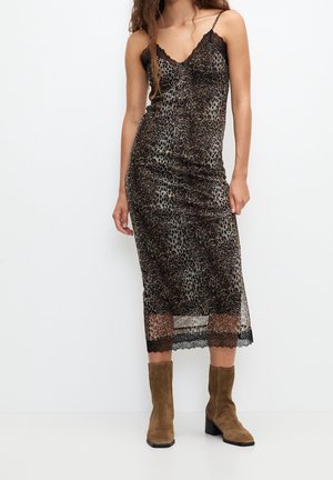 Woman wearing a sleeveless, leopard-print midi dress with lace trim and brown suede ankle boots, standing against a plain white background.